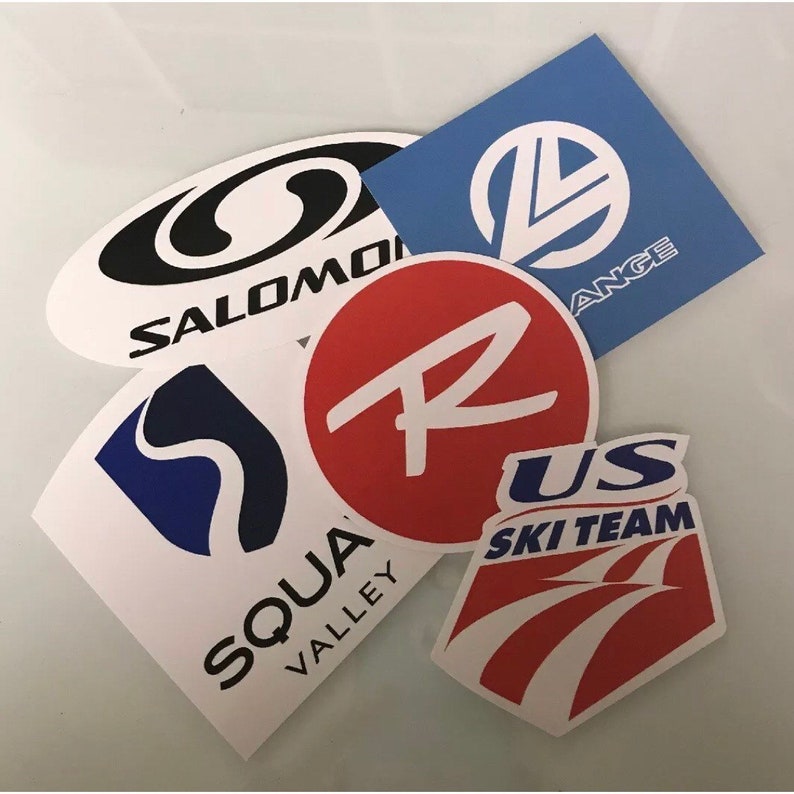 May include: Assortment of five white stickers with various logos and text. The logos include a black and white Salomon logo, a blue and white Squaw Valley logo, a red circle with a white letter R, a blue circle with a white letter L and the text "Lange", and a red and white US Ski Team logo.