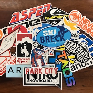 May include: A collection of colorful stickers featuring various ski resort and snowboard company logos, including "Aspen", "Telluride", "Ski Breck", "Park City", "Never Summer", and "Quicksilver".