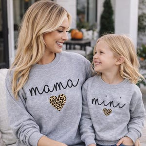 Può includere: Due felpe grigie abbinate, una con la scritta "mama" e l'altra con la scritta "mini" in corsivo nero, ciascuna con un cuore con stampa leopardata. Le felpe sono indossate con jeans blu e pantaloncini di jeans.