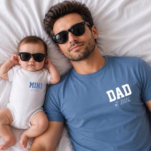 Könnte beinhalten: Ein blaues T-Shirt mit dem Wort "DAD" und dem Jahr "2026" in Weiß aufgedruckt. Ein Baby in einem weißen Body mit dem Wort "MINI" in Blau aufgedruckt. Beide tragen schwarze Sonnenbrillen und liegen auf einem weißen Laken.