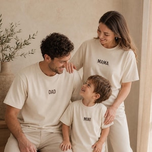 Könnte beinhalten: Drei passende cremefarbene Kurzarm-T-Shirts. Ein T-Shirt trägt den Schriftzug DAD in Dunkelbraun, ein weiteres MAMA in Dunkelbraun und das dritte MINI in Dunkelbraun. Jedes T-Shirt hat einen Rundhalsausschnitt und eine lockere Passform.