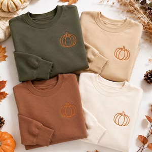 Peut inclure: Quatre sweat-shirts à col rond en vert olive, beige, marron et crème, chacun arborant une petite citrouille brodée orange sur la poitrine gauche. Les vêtements à manches longues sont disposés en grille sur une surface blanche.