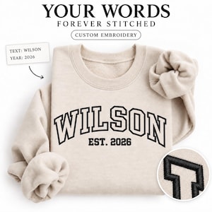 Könnte beinhalten: Ein beigefarbenes Sweatshirt mit Rundhalsausschnitt und schwarzer Stickerei, die WILSON und EST. 2026 in einer College-Schriftart zeigt. Das Set enthält ein passendes beiges Haargummi. Textmarkierungen zeigen Optionen für individuelle Stickereien.