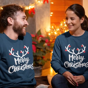 May include: Two people wearing matching navy blue sweatshirts with a white antler design and the words "Merry Christmas" in a script font. The antlers have red ball ornaments. The background includes Christmas lights.