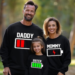 May include: A family of three wearing black sweatshirts with a battery graphic and the words "Daddy", "Mommy", and "Sister" on them. The battery graphic is filled with red for the "Daddy" shirt, a lightning bolt for the "Mommy" shirt, and green for the "Sister" shirt.