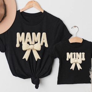 May include: Black t-shirts featuring the words "MAMA" and "MINI" in a cream-coloured, textured font, each with a matching bow design. The shirts are displayed on wooden hangers against a white backdrop.