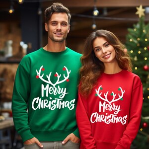 May include: Green and red crewneck sweatshirts with the text "Merry Christmas" and a white antler design. The sweatshirts are worn by two people in front of a Christmas tree.