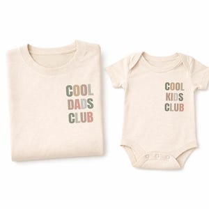 以下が含まれることがあります： クリーム色のシャツ2枚。1枚は折りたたまれ、もう1枚はベビーボディスーツ。どちらも「COOL DADS CLUB」と「COOL KIDS CLUB」の文字が、くすんだ緑、茶色、ピンクの文字で重ねてデザインされています。柔らかい生地で作られているようです。