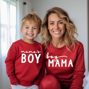 May include: Matching red sweatshirts with white text that reads "mama's BOY" and "boy MAMA". A woman and a child are wearing the sweatshirts and smiling at the camera. The sweatshirts are a solid red color.