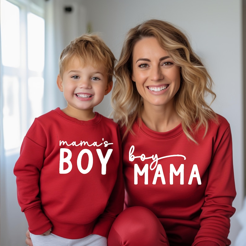 May include: Matching red sweatshirts with white text that reads "mama's BOY" and "boy MAMA". A woman and a child are wearing the sweatshirts and smiling at the camera. The sweatshirts are a solid red color.