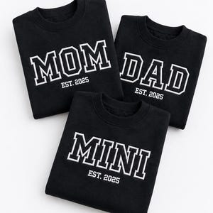 Pode incluir: Três camisolas pretas de gola redonda com letras bordadas a branco. Uma camisola apresenta o texto MOM EST. 2025, outra DAD EST. 2025 e a terceira MINI EST. 2025. O texto está numa fonte a negrito de estilo universitário.