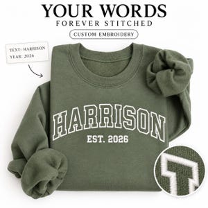 Könnte beinhalten: Ein olivgrüner Rundhals-Pullover mit weißem Sticktext, der HARRISON EST. 2026 in einer College-Schriftart zeigt. Das Design enthält einen kreisförmigen Ausschnitt, der die detaillierte weiße Fadenstickerei des Buchstabens H zeigt.