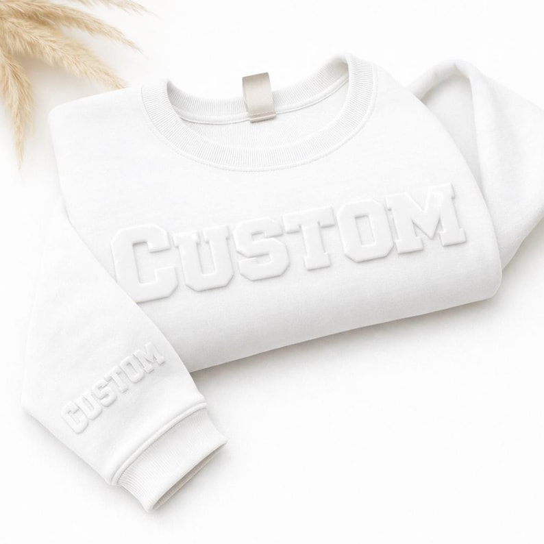 以下が含まれることがあります： 胸元にCUSTOMという文字が浮き彫りになった白い長袖のスウェットシャツ。同じCUSTOMという文字が、リブ編みの袖口に近い左袖の下部にも小さく浮き彫りにされています。