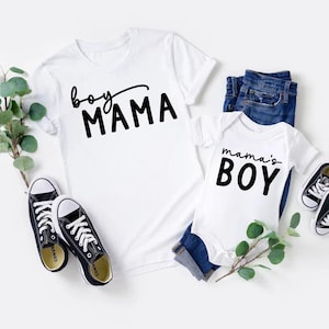 May include: White t-shirt with "boy MAMA" in black script and a white baby onesie with "mama's BOY" in black. Includes blue jeans and black canvas sneakers. Greenery accents the display.