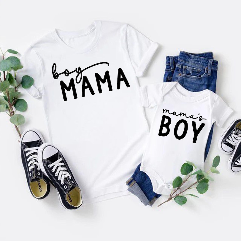May include: White t-shirt with "boy MAMA" in black script and a white baby onesie with "mama's BOY" in black. Includes blue jeans and black canvas sneakers. Greenery accents the display.