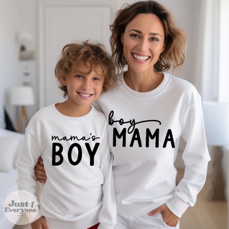 May include: White sweatshirts with black text. One sweatshirt says "mama's BOY", and the other says "boy MAMA". The sweatshirts are long-sleeved and worn by a mother and child. The image is taken indoors.