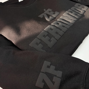 以下が含まれることがあります： 胸元にマットブラックの立体的な質感で「Z&Eacute; FERRINHOS」の文字が施された黒の長袖スウェットシャツ。袖には同じマットブラックの立体的な素材で「ZF」の文字があしらわれており、全体的にモノクロームな外観に仕上がっています。