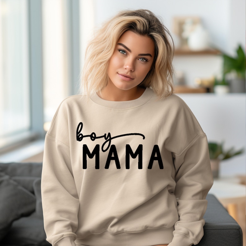 May include: A beige sweatshirt with the words "boy MAMA" printed in black. The word "boy" is in a cursive font, and "MAMA" is in a bold, block font. The sweatshirt is a neutral color.