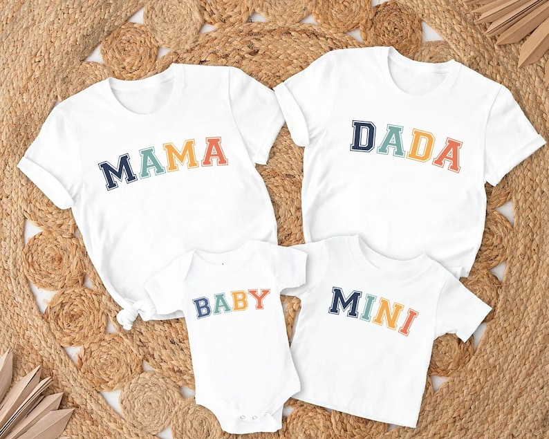 Op de afbeelding: Vier witte T-shirts met kleurrijke tekst. De shirts zeggen "Mama", "Dada", "Baby" en "Mini".
