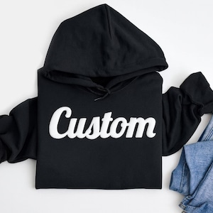 以下が含まれることがあります： 胸元に白い筆記体でCustomという文字がプリントされた黒いフード付きスウェットシャツと、その横にきれいに折りたたまれた青いデニムパンツが、無地の白い表面の上に配置されています。