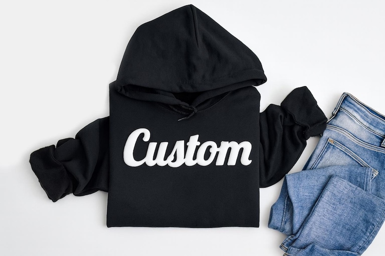 以下が含まれることがあります： 胸元に白い筆記体でCustomという文字がプリントされた黒いフード付きスウェットシャツと、その横にきれいに折りたたまれた青いデニムパンツが、無地の白い表面の上に配置されています。