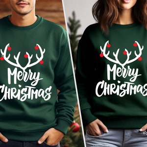 May include: Two dark green sweatshirts with the text "Merry Christmas" in white script. Above the text is a white antler design with red ornaments. The sweatshirts are a festive holiday theme.