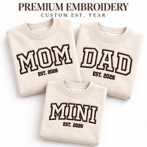 Peut inclure: Trois sweat-shirts à col rond de couleur crème avec du texte brodé marron foncé. Les sweat-shirts affichent les mots MOM, DAD et MINI dans une police universitaire, chacun accompagné du texte EST. 2026. Le texte en haut indique PREMIUM EMBROIDERY CUSTOM EST. YEAR.