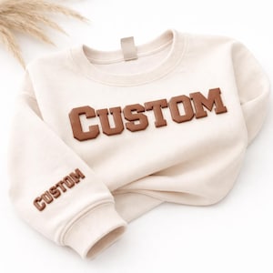以下が含まれることがあります： 胸元に茶色の立体的なブロック体で「CUSTOM」という文字が施された、クリーム色のクルーネックのスウェットシャツ。左袖の下部にも、より小さな茶色の立体的なブロック体で「CUSTOM」という文字がデザインされています。