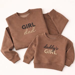Op de afbeelding: Twee bruine sweatshirts, één voor volwassenen en één voor kinderen, met bijpassende teksten. Het sweatshirt voor volwassenen zegt "GIRL dad" in zwarte en gouden borduursels. Het sweatshirt voor kinderen zegt "daddy's GIRL" in zwarte en gouden borduursels.