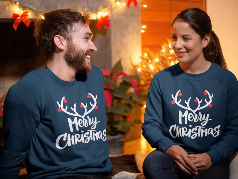 May include: Two people wearing matching navy blue sweatshirts with a white antler design and the words "Merry Christmas" in a script font. The antlers have red ball ornaments. The background includes Christmas lights.