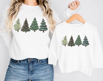May include: White sweatshirts featuring a row of illustrated Christmas trees. The trees are green with various colored lights and star or snowflake toppers. One sweatshirt is adult-sized, and the other is child-sized, both with long sleeves.