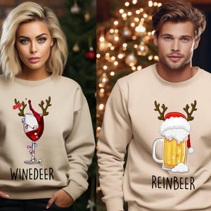 Könnte beinhalten: Beige Sweatshirts mit festlichen Grafiken. Eines zeigt ein Weinglas mit Geweih und dem Wort "WINEDEER". Das andere zeigt einen Bierkrug mit Weihnachtsmütze und dem Wort "REINBEER". Ideal für Weihnachten.