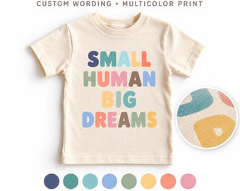 以下が含まれることがあります： クリーム色の半袖Tシャツで、丸みを帯びたマルチカラーのフォントで「SMALL HUMAN BIG DREAMS」という文字がプリントされています。円形のインセットには生地の質感のクローズアップが表示されています。Tシャツの下には8つの色付きの円とテキストがあります。