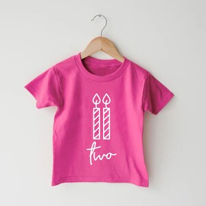 Puede incluir: Camiseta rosa de manga corta con un gr&aacute;fico blanco de dos velas de cumplea&ntilde;os y la palabra "dos" escrita en letra cursiva.