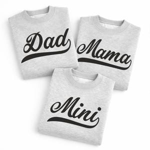 Op de afbeelding: Drie lichtgrijze sweatshirts met de woorden "Dad", "Mama" en "Mini" in zwarte letters. De sweatshirts liggen op een witte ondergrond.