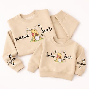 Könnte beinhalten: Beige Sweatshirts mit Winnie-the-Pooh-Grafiken. Eines der Sweatshirts trägt die Aufschrift "mama bear", das andere "baby bear". Beide zeigen einen Cartoon-Bären mit Honigtopf und Bienen. Der Ärmel von "mama bear" trägt die Aufschrift "Sophia".