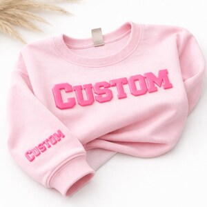 以下が含まれることがあります： 胸元に大きなダークピンクの立体的なバーシティスタイルの文字で「CUSTOM」と書かれ、左袖の下部にも小さなダークピンクの立体的な文字で「CUSTOM」と書かれた、ライトピンクのクルーネックスウェットシャツ。
