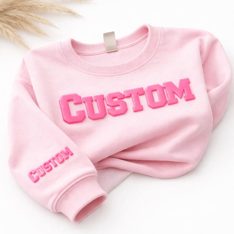 以下が含まれることがあります： 胸元に大きなダークピンクの立体的なバーシティスタイルの文字で「CUSTOM」と書かれ、左袖の下部にも小さなダークピンクの立体的な文字で「CUSTOM」と書かれた、ライトピンクのクルーネックスウェットシャツ。