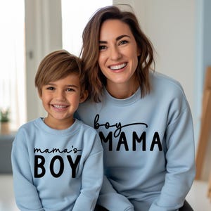 May include: A mother and son wearing matching light blue sweatshirts. The mother's sweatshirt says "boy MAMA" in black script and block letters. The son's sweatshirt says "mama's BOY" in black block letters.