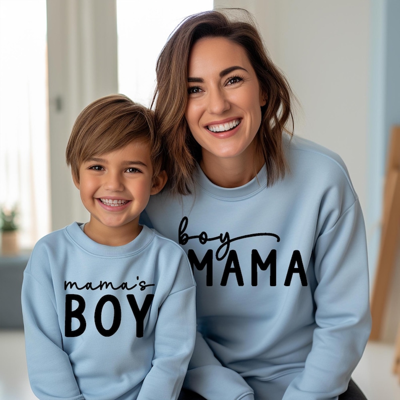 May include: A mother and son wearing matching light blue sweatshirts. The mother's sweatshirt says "boy MAMA" in black script and block letters. The son's sweatshirt says "mama's BOY" in black block letters.