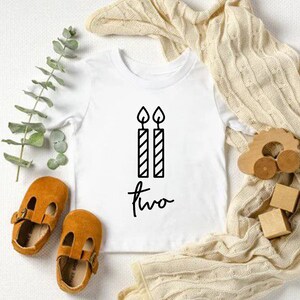 Puede incluir: Un body blanco para beb&eacute; con texto negro que dice "dos" y un gr&aacute;fico de dos velas con llamas.