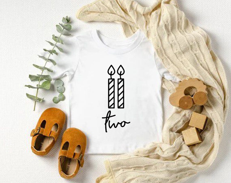 Puede incluir: Un body blanco para beb&eacute; con texto negro que dice "dos" y un gr&aacute;fico de dos velas con llamas.
