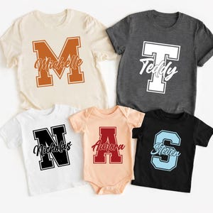 Peut inclure: Cinq t-shirts personnalisés et un body. Les t-shirts sont crème, anthracite, blancs et noirs. Le body est pêche. Chaque vêtement présente une grande lettre avec un nom en dessous en écriture script. Les lettres sont orange, blanches, noires, rouges et bleu clair.