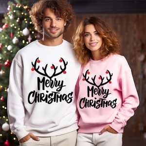 May include: White and pink sweatshirts with a black antler design and the words "Merry Christmas" in a script font. The antlers have red ornaments. The sweatshirts are part of a matching set.