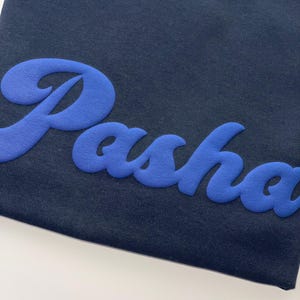 以下が含まれることがあります： 胸元に明るい青色の立体的な質感のある筆記体で「Pasha」という文字がデザインされた、ダークネイビーのTシャツ。文字は柔らかいマットな仕上がりで、暗い色の生地に対して立体的に浮かび上がっています。