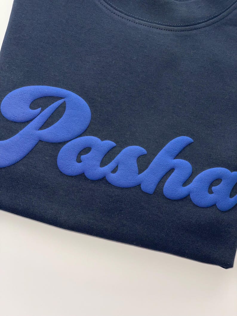 以下が含まれることがあります： 胸元に明るい青色の立体的な質感のある筆記体で「Pasha」という文字がデザインされた、ダークネイビーのTシャツ。文字は柔らかいマットな仕上がりで、暗い色の生地に対して立体的に浮かび上がっています。