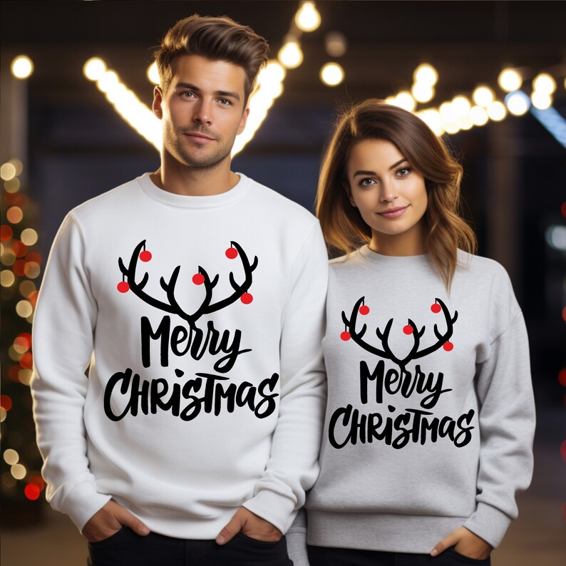 May include: Two people wearing matching Christmas sweatshirts. One sweatshirt is white, the other is gray. Both have a black antler design with red ornaments and the words "Merry Christmas" in black script.