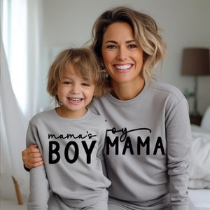 May include: A mother and child wearing matching gray sweatshirts. The child's sweatshirt says "mama's BOY" in black script, while the mother's says "joy MAMA". The image captures a moment of joy and connection.
