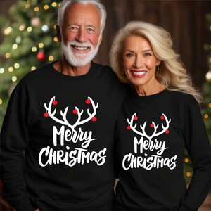 May include: Two people wearing matching black sweatshirts. Each sweatshirt has a white antler design with red ornaments and the words "Merry Christmas" in white script. The background is a blurred Christmas tree.