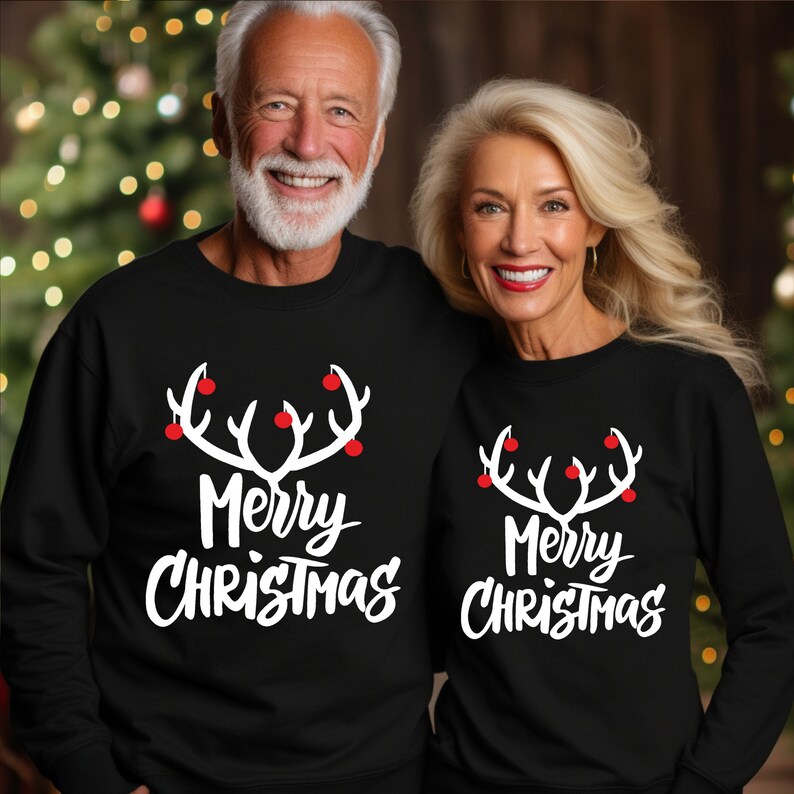 May include: Two people wearing matching black sweatshirts. Each sweatshirt has a white antler design with red ornaments and the words "Merry Christmas" in white script. The background is a blurred Christmas tree.